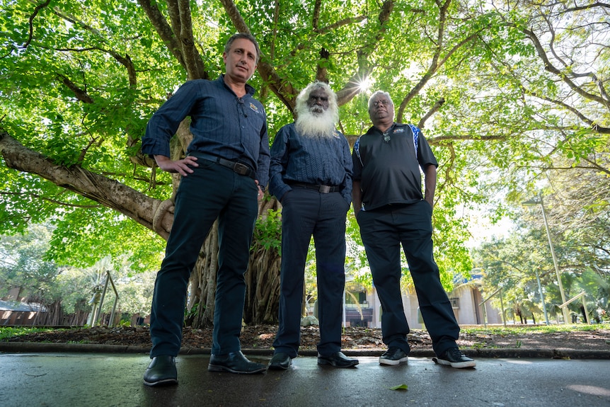 Three men part of the East Arnhem Council standing under a tree, looking serious, on a sunny day.