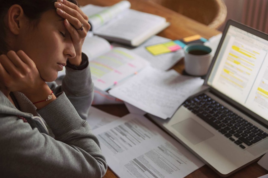 Young woman with burnout syndrome holds her head as she sits in front of a computer which is surrounded by papers.