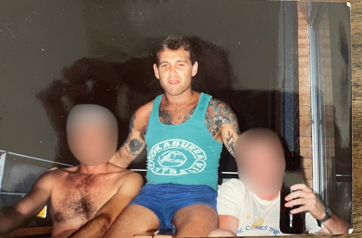 A man with tattoos in a blue singlet with his arms around two other men whose faces are blurred.
