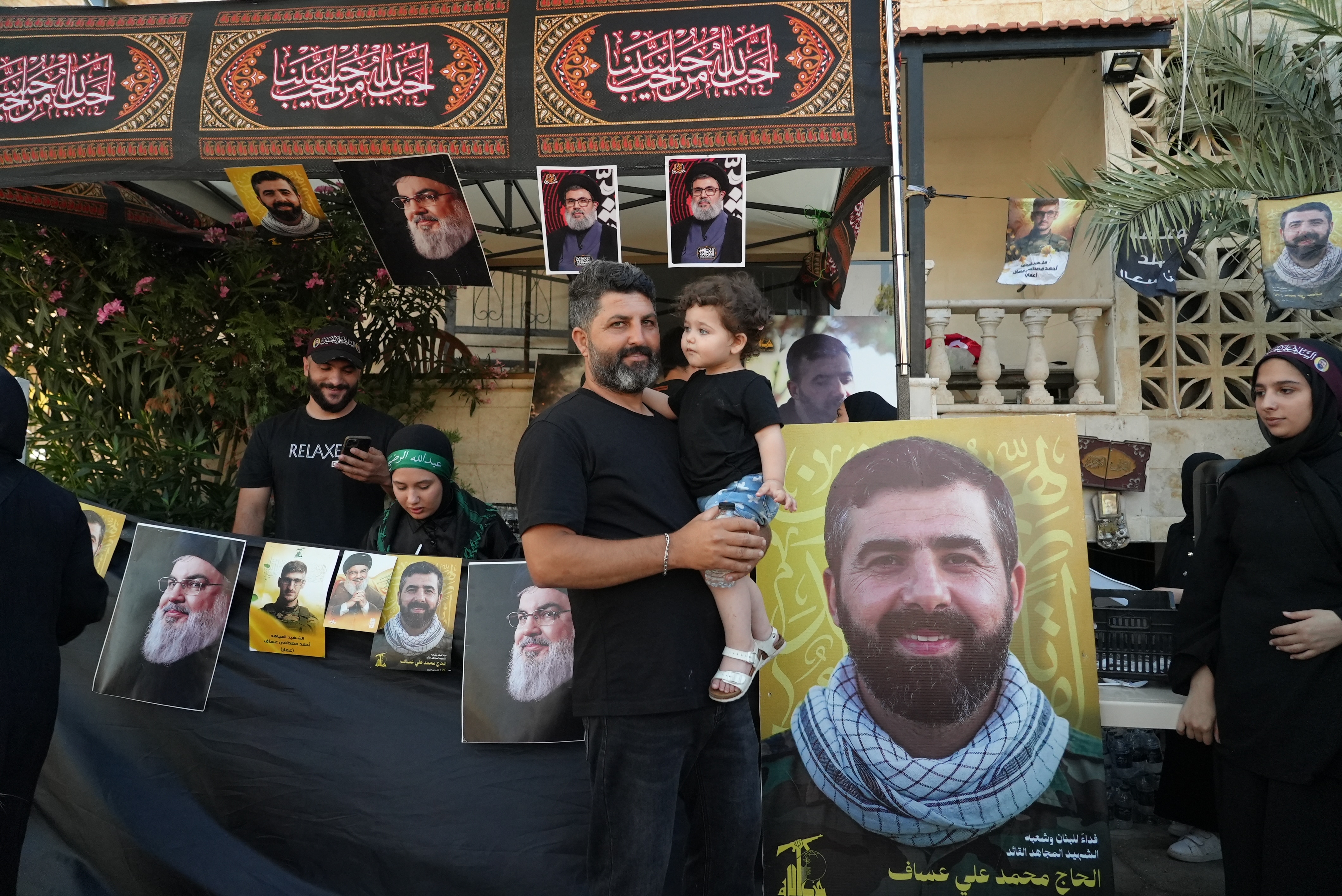 A man with a greying beard holds up a child. Behind them are pictures of Hezbollah fighters and leaders.