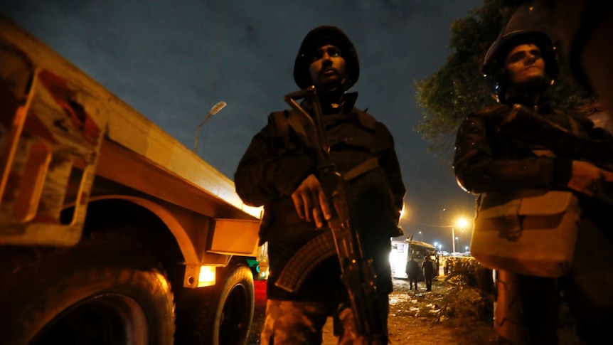 Two armed men standing by a truck at night, with their faces partially-lit by street lights.