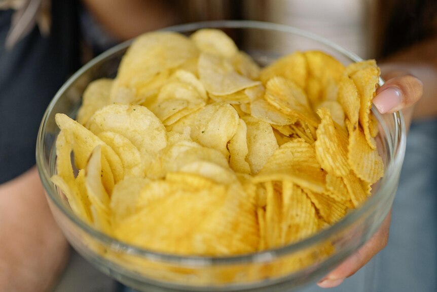 A woman's hand takes a potato chip from a clear bowl she is holding, filled with them.