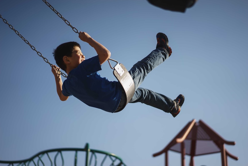 A boy swings high on a playground swing, sun shining on his smiling face.