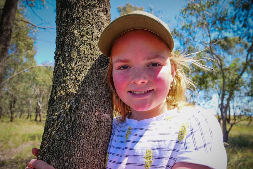 A young girl in a cap stands hugging a tree.