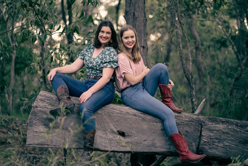 Two women sit on a large fallen tree trunk, back to back. They're smiling at the camera