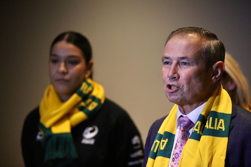 A man in a suit with a yellow and gold scarf speaks.