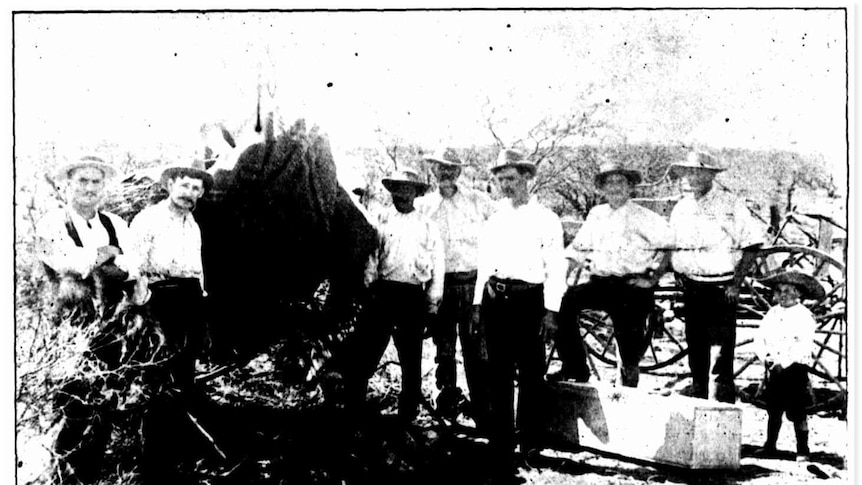 An historic photo shows men and a boy standing around a mine shaft where a dead body has been brought to the surface