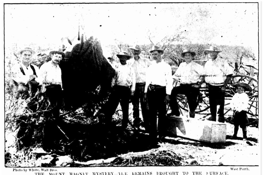 An historic photo shows men and a boy standing around a mine shaft where a body has been brought to the surface.