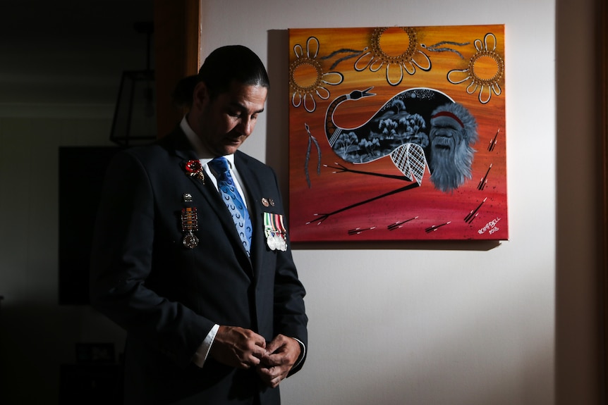 A man in a suit with medals stands in front of an Indigenous artwork.