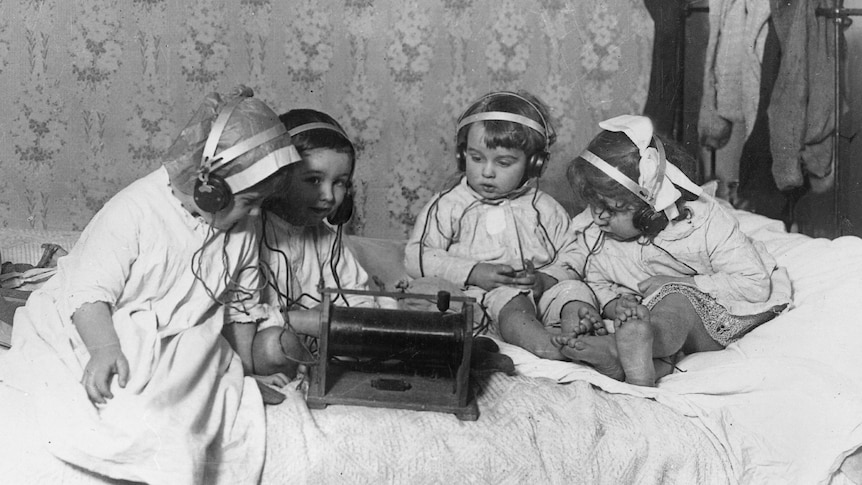 a group of children sitting on a bed and listening to the radio on headphones circa 1930