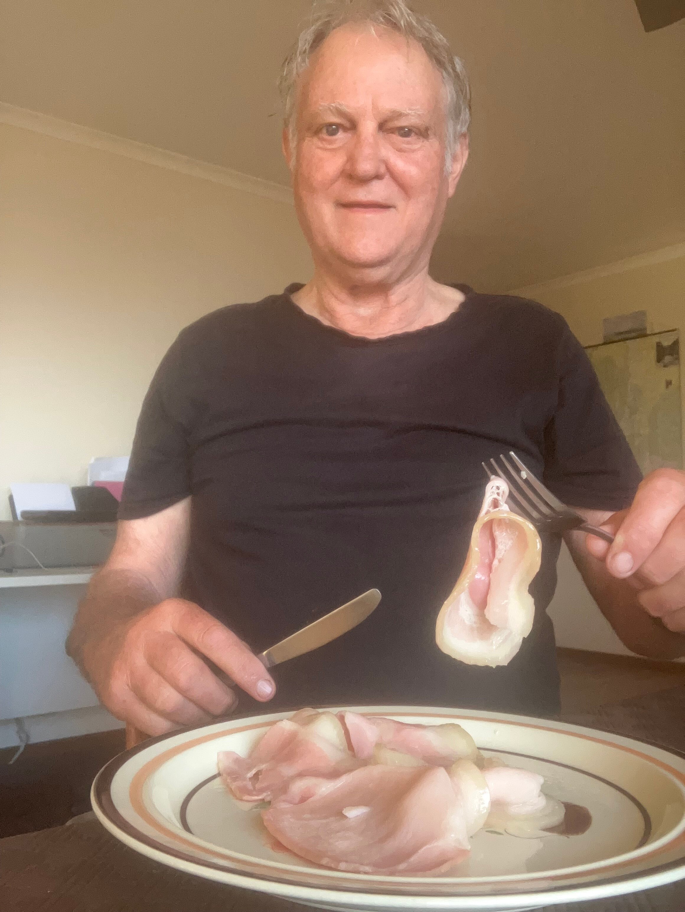 A person seated at a table with a plate of bacon in front of him, holding up a rash on a fork.