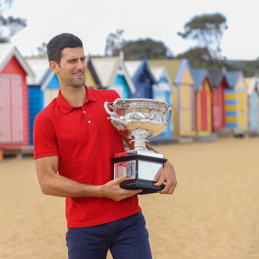 Novak Djokovic stands in front of some brightly coloured beach huts in a red polo shirt, holding the Australian Open trophy