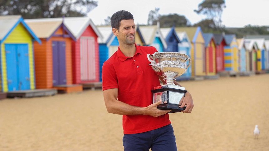 Novak Djokovic stands in front of some brightly coloured beach huts in a red polo shirt, holding the Australian Open trophy