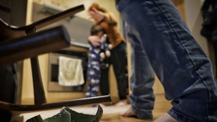 A woman holds her child and covers her face in the background, a broken cup on the floor and a man's legs in the foreground