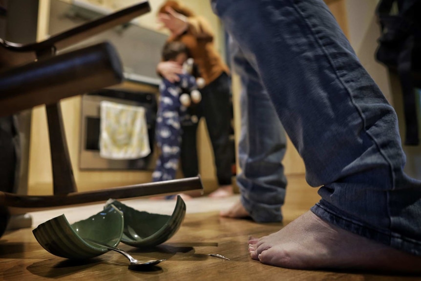 A woman holds her child and covers her face in the background, a broken cup on the floor and a man's legs in the foreground