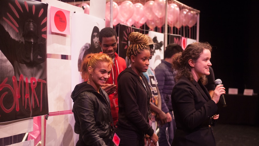 2 women and 3 men stand on a stage smiling behind founder of the project Rachel Barnard who is smiling speaking into microphone