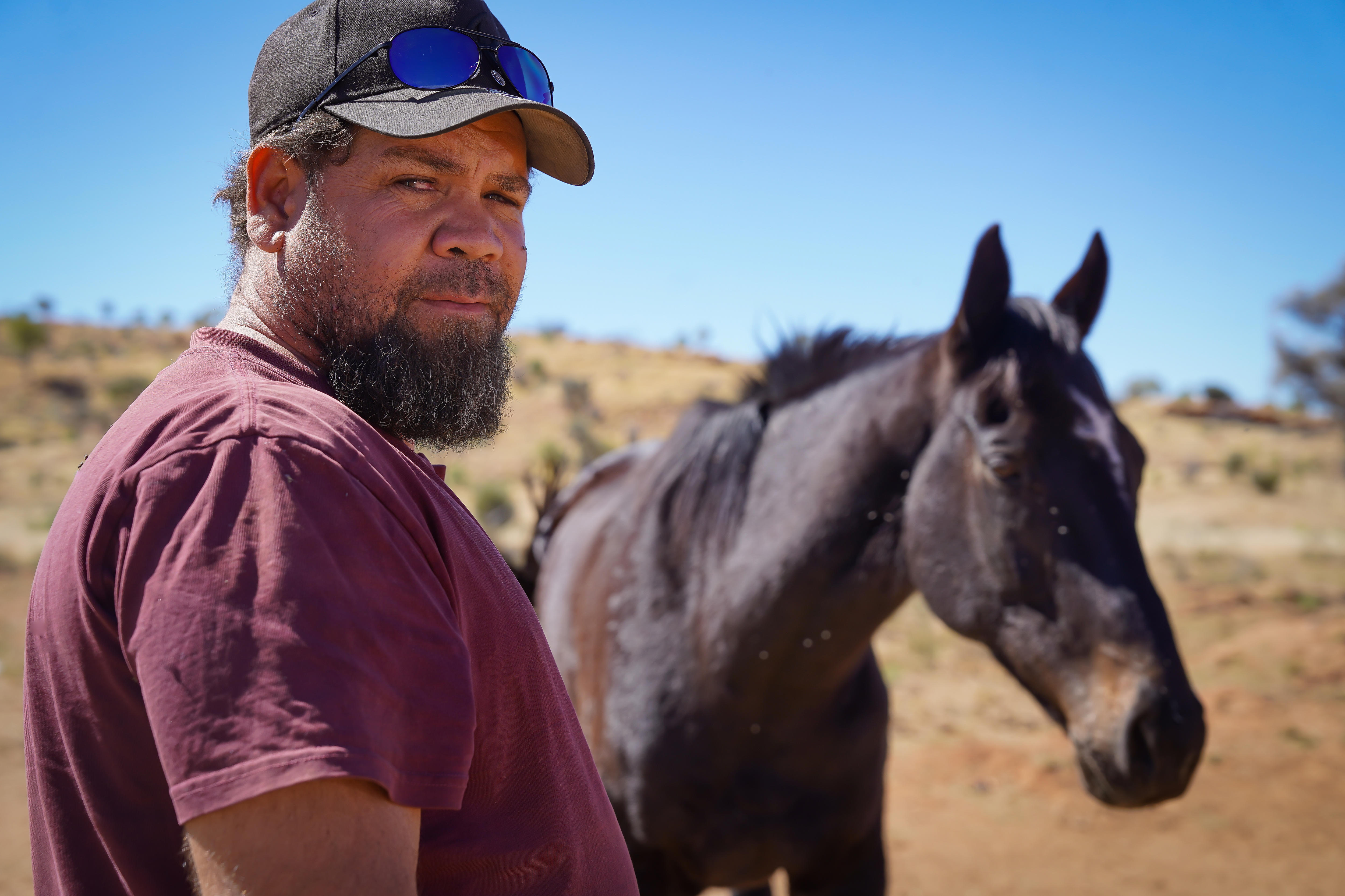 an aboriginal man with a beard wearing a cap, standing next to a horse