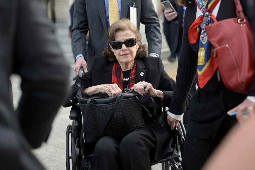 Dianne Feinstein is in a wheelchair. She wears sunglasses and holds a black bag.