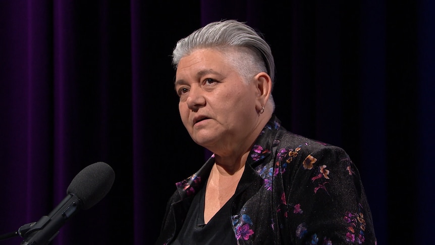 Middle-aged woman with slicked back short grey hair wears black blazer with purple flowers on it and speaks into mic