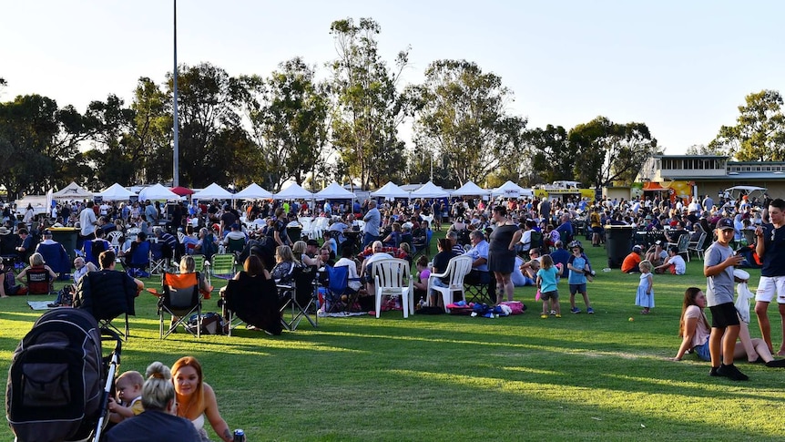 A large crowd of people is spread across a sporting oval. There are white tents in the background.