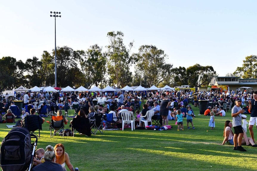 A large crowd of people is spread across a sporting oval. There are white tents in the background.