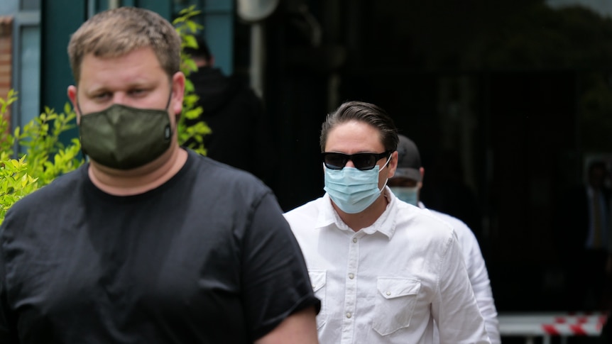 A man in a black shirt and green face mask and a man with dark hair and sunglasses in a white shirt