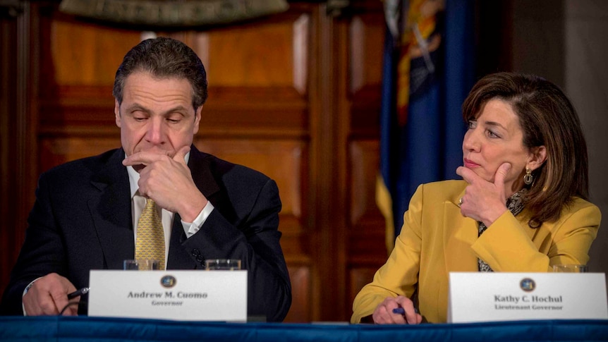 A woman holds her right hand across her face as she looks at a suited man sitting next to her, who is gazing downwards.