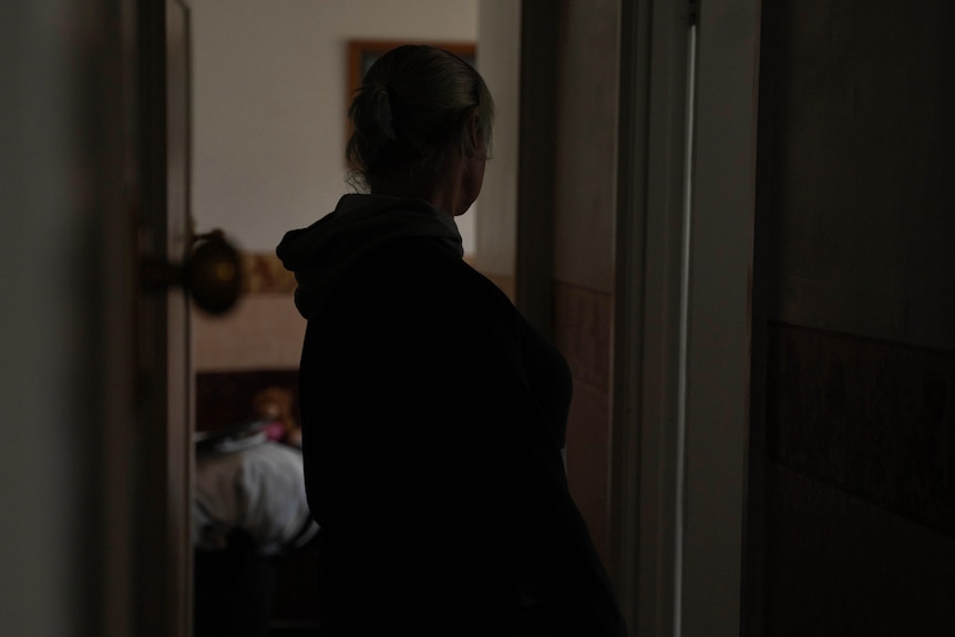 A woman stands in profile in a darkened corridor with an open bedroom door behind her 