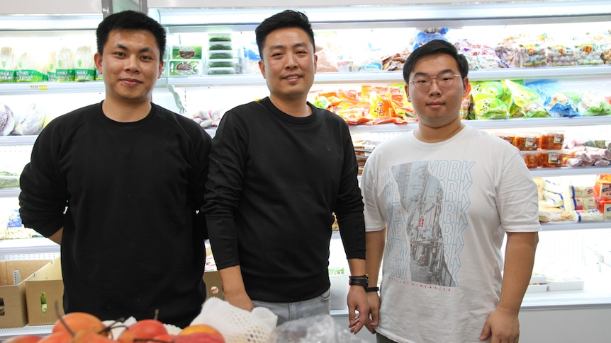 Three Chinese men stand in front of a well stocked fridge.