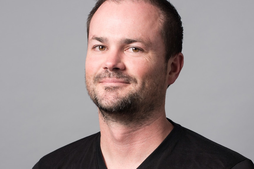 A headshot of a man with short hair and a light beard, wearing a dark shirt.
