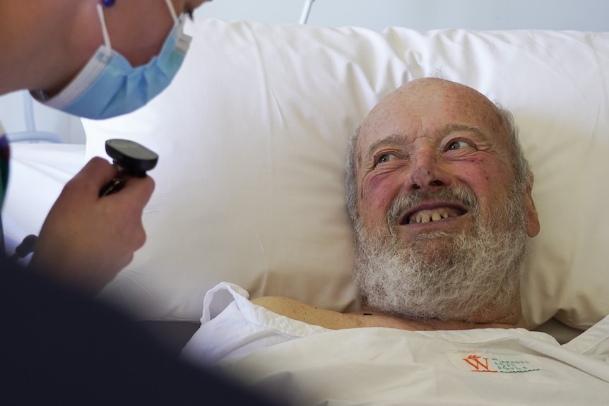 A photo of a man in a hospital bed smiling at his doctor who is wearing a facemask