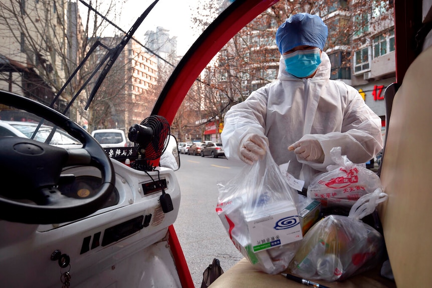 From inside a small van, you see a person in a hazmat suit, face mask and hair net place plastic bags of medicine on a seat.