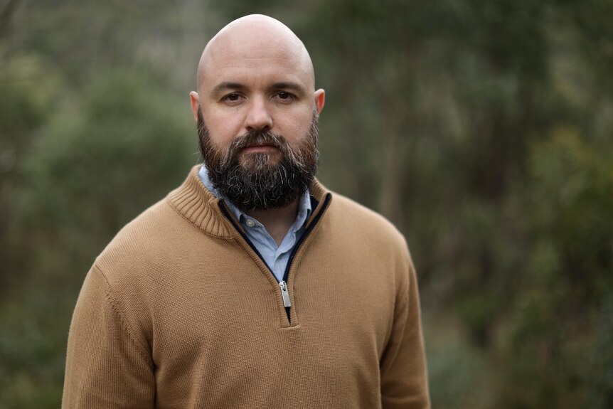 A bald man with a beard stands outside, greenery behind him, looking directly at the camera with a neutral expression.