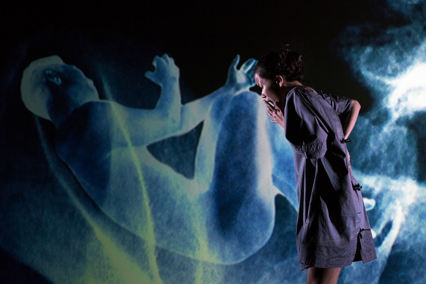 An Asian Australian woman in a blue dress looks distressed, as she looks down, a blue image of a man's body projected behind her