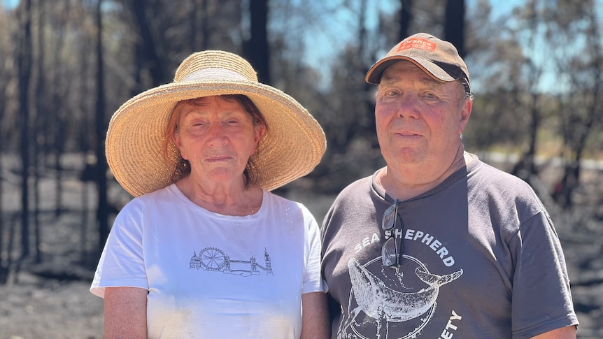 A man and a woman wearing hats on a sunny day with burnt trees and charred ground behind them.