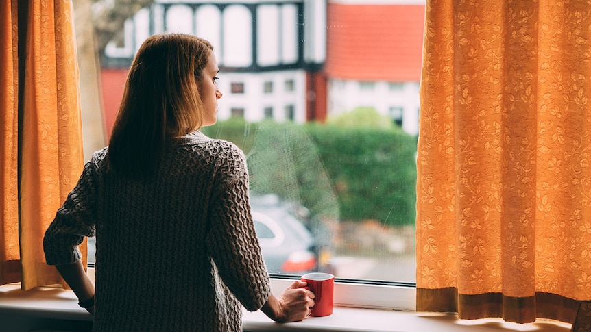 Woman looking out of window onto street, with orange curtains beside her framing window.