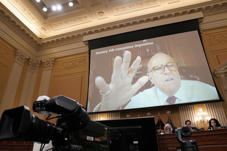 Rudy Giuliani, his hand extended, is seen on a screen inside an ornate room with cameras and people seated at desks