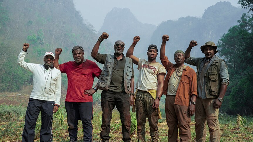 On hazy day in mountainous jungle six men  with serious expressions wear hiking attire and stand with raised right fists.