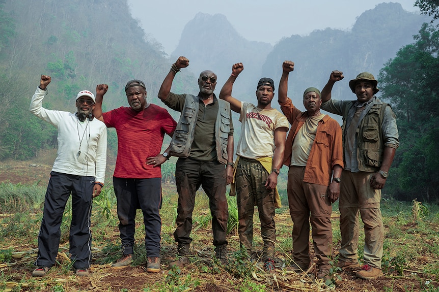 On hazy day in mountainous jungle six men with serious expressions wear hiking attire and stand with raised right fists.