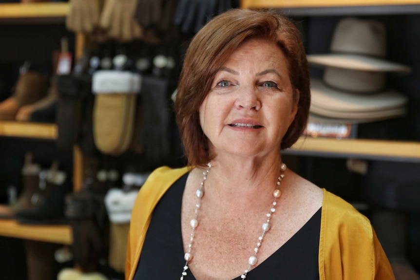 A head and shoulders shot of an older woman standing in a shop in front of hats and gloves on shelves.