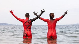 Two nude people stand in a river with red painted bodies advertising Dark MOFO's nude swim.