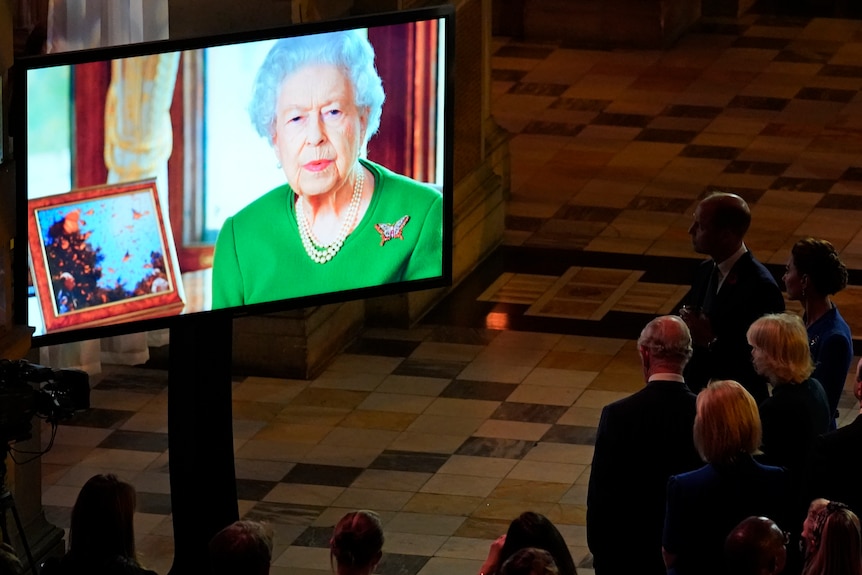 Queen in an green dress on a screen watched by others.