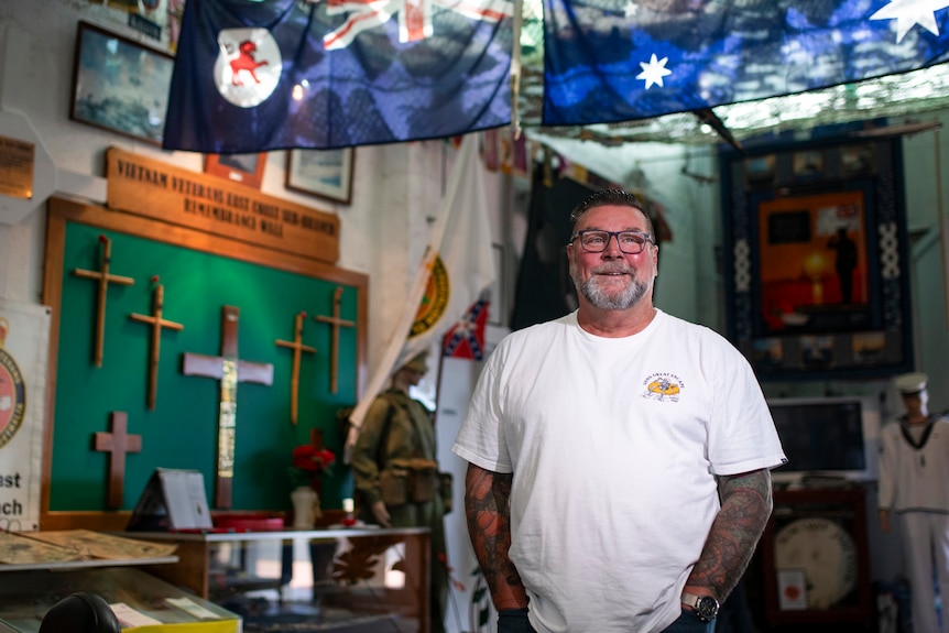 A man in a white t-shirt and navy-framed glasses stands in a room filled with ANZAC memorabilia and flags hanging above him.