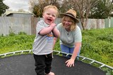 Small boy on trampoline with woman wearing hat behind him