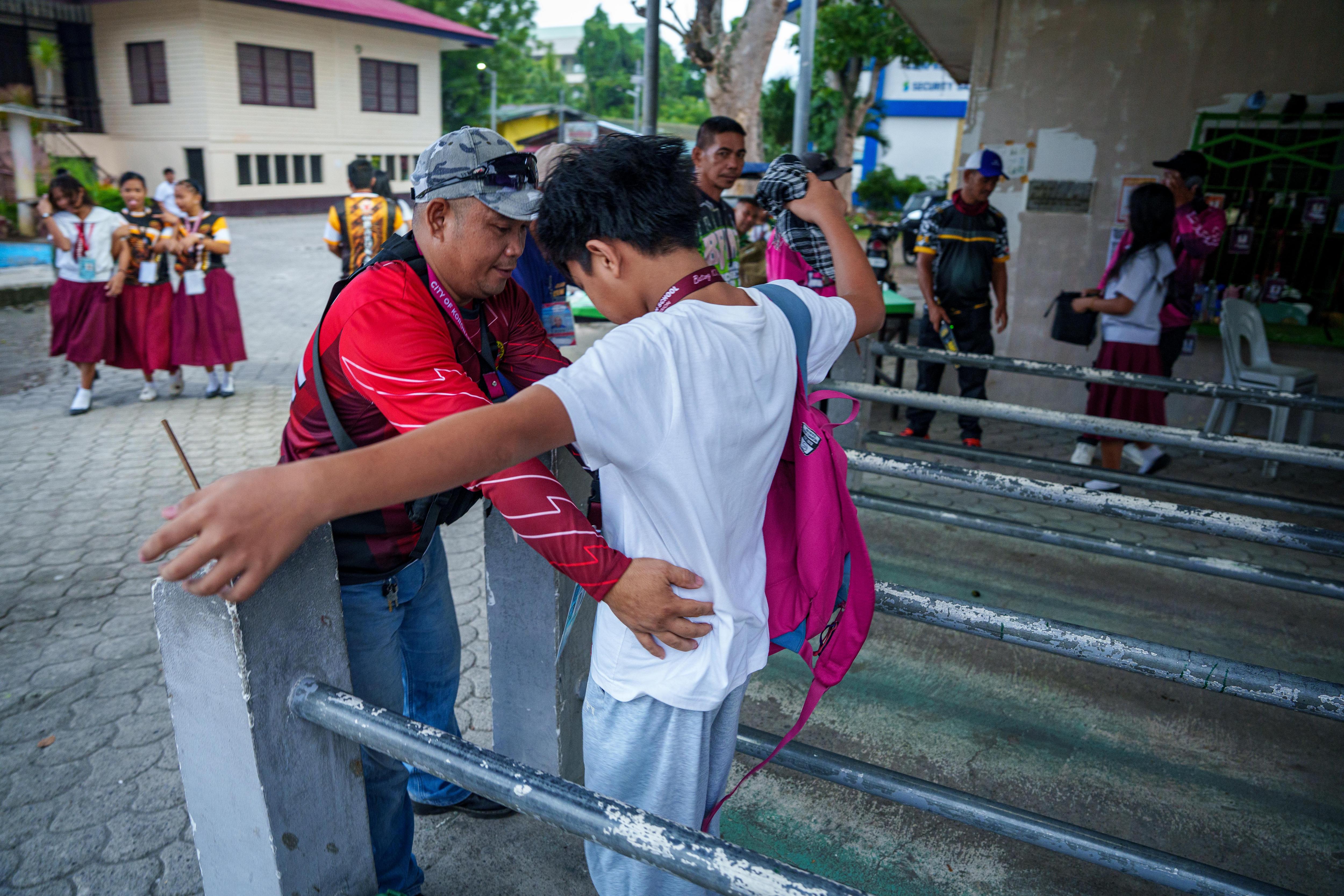 A young Filipino schoolboy outstretches his arms while being patted down by a security guard.