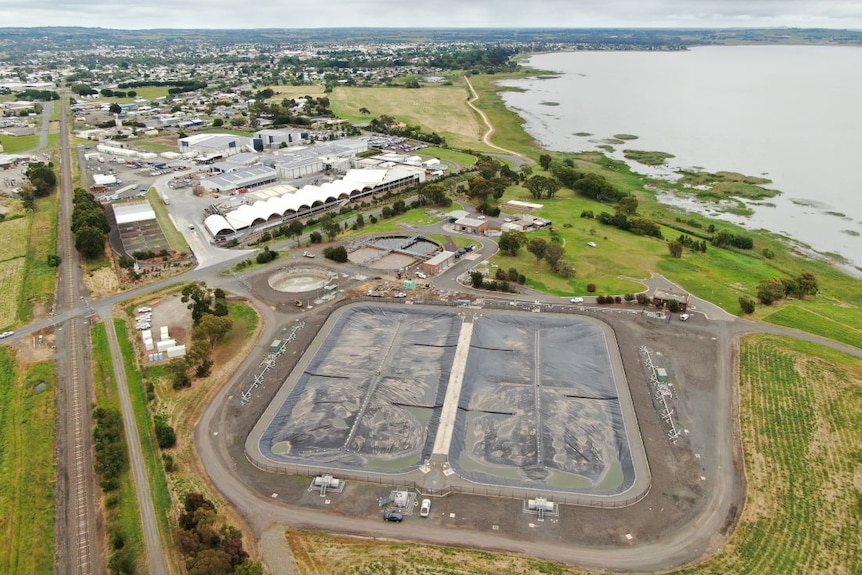 An aerial shot of four large rectangular lagoons covered in black plastic, at an industrial site beside a lake.