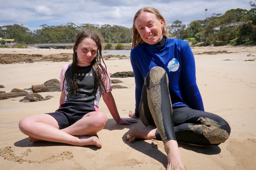 A girl and a woman sit on a beach in wetsuits looking at the camera, smiling. 