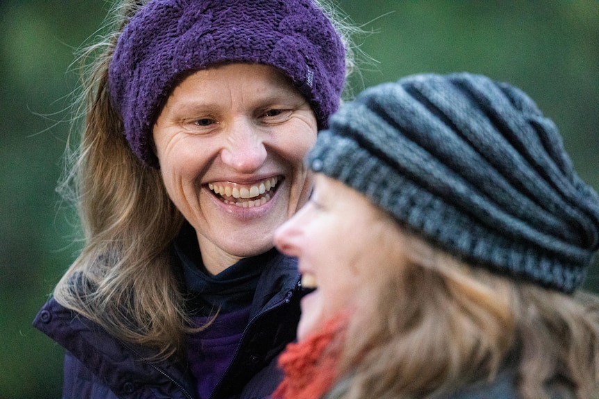 A woman smiling at another woman. She is wearing a purple knitted beanie and a jacket.