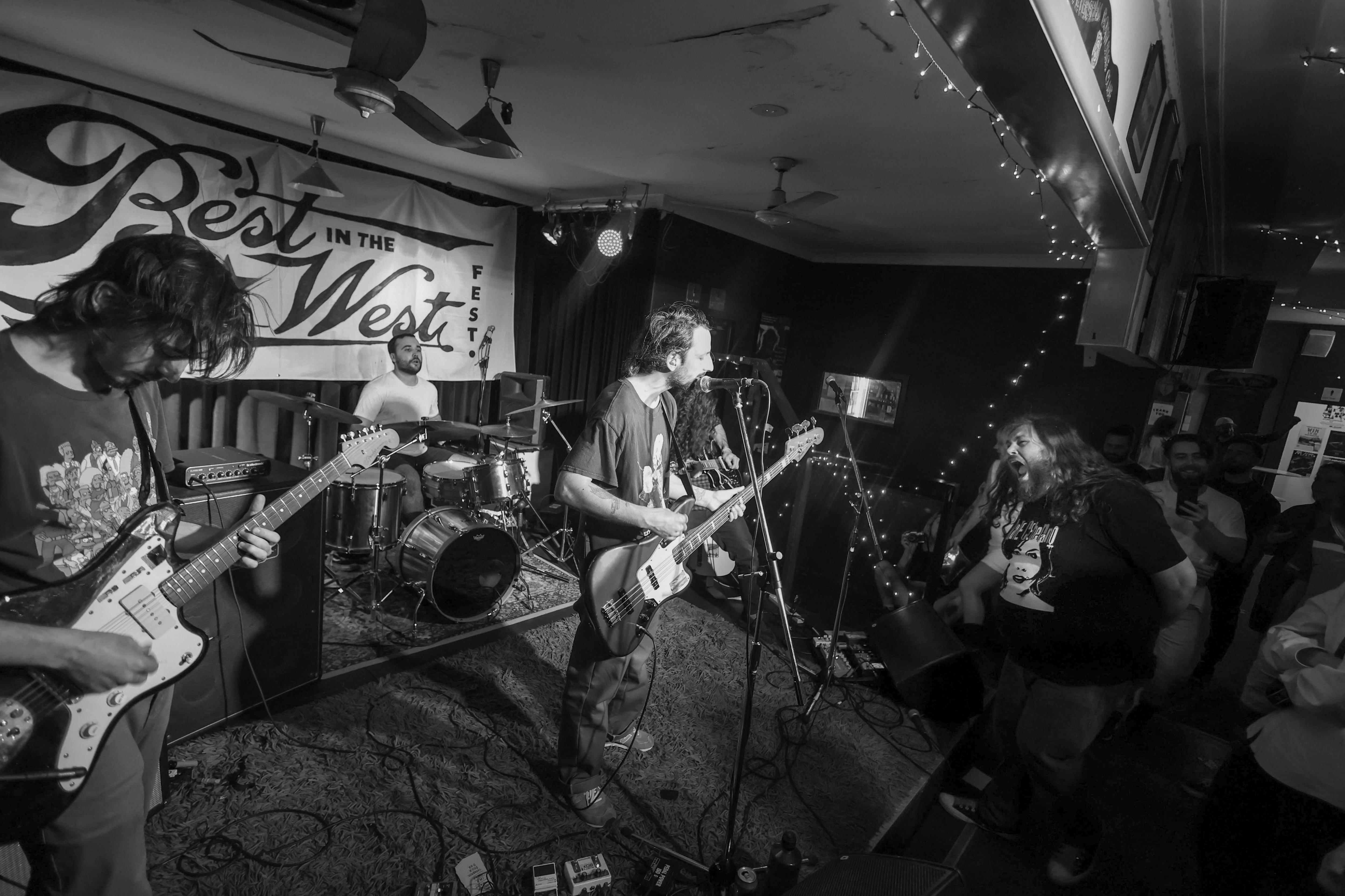 A man with long hair beard stands in a small room watching a three-piece all-male band comprising guitar, drums and bass. 