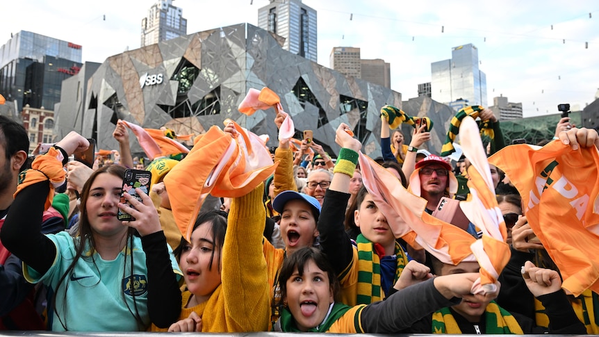 A cheering crowd holding green and gold banners and flags celebrate behind a metal fence in Melbourne's Federation Square.